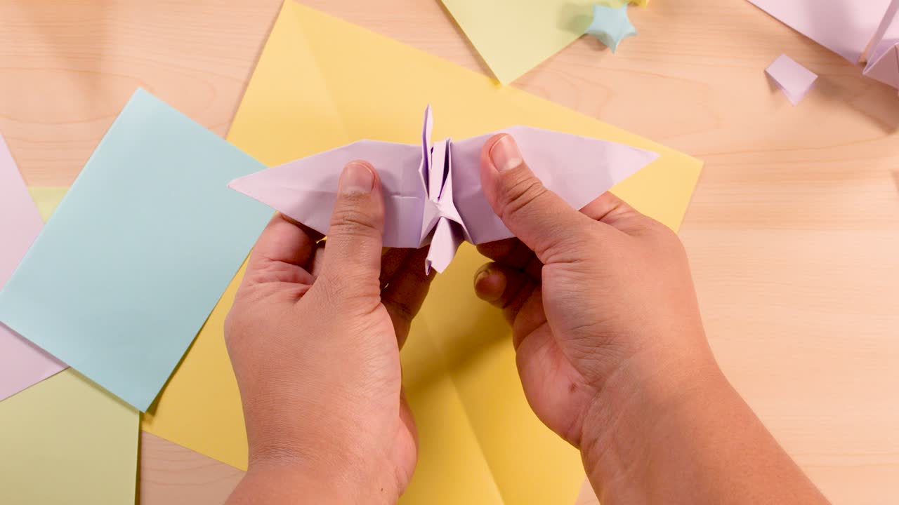 Person folds paper origami bird on pastel paper background, overhead shot, soft even lighting
