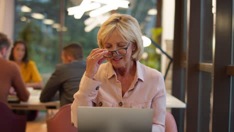 mature businesswoman working on laptop at desk in office pausing to look out of window