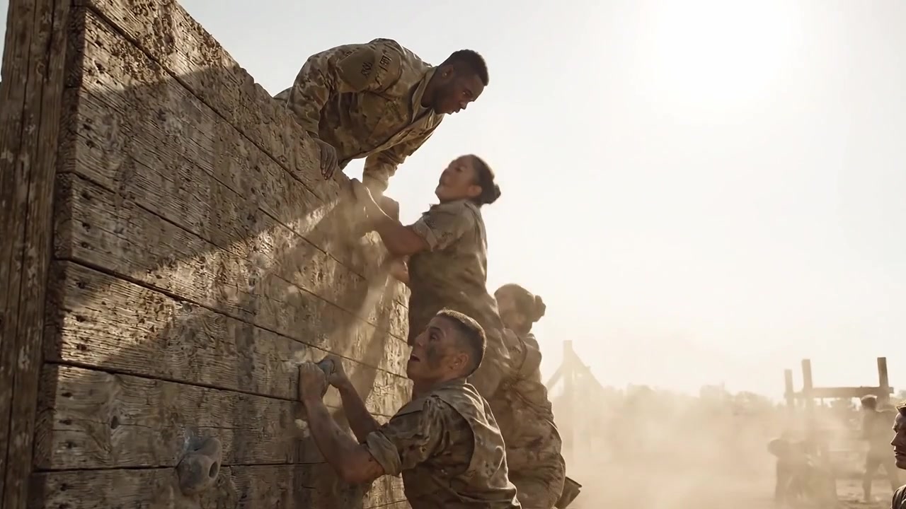 Un gruppo di soldati affronta un percorso a ostacoli durante l'addestramento, mostrando lavoro di squadra e determinazione mentre collaborano per scalare un muro di legno in condizioni polverose.