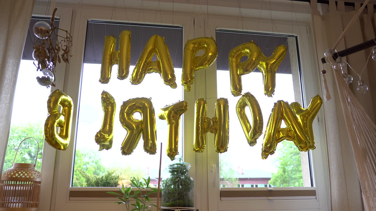 letras mayúsculas feliz cumpleaños de globos dorados moviéndose frente a la ventana del apartamento
