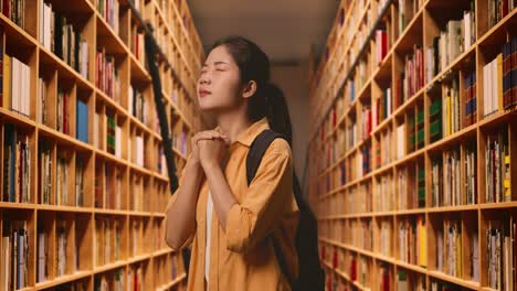 vista lateral de una estudiante asiática con una mochila cerrando los ojos reza por algo mientras está de pie en la biblioteca