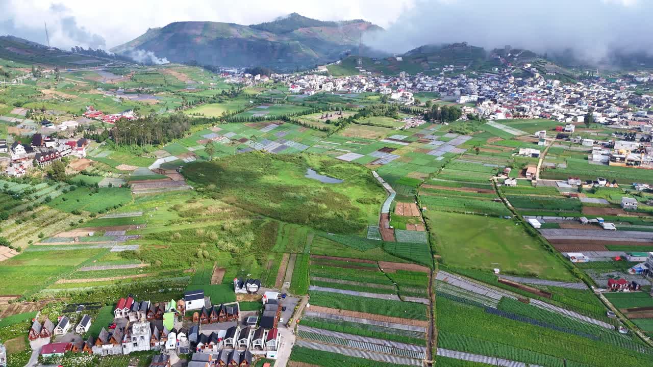 Aerial drone view of a scenic mountain valley with lush green farmland, small houses, and a town surrounded by misty hills. Peaceful rural landscape on a bright sunny morning in a highland region