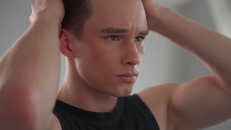 close up of satisfied young man in black vest styling hair with hands, content expression, blurred mirror reflection in background, soft daylight illuminating skin texture in bright minimalist studio