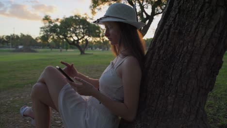 Hermosa-Joven-Con-Sombrero-Apoyado-En-Un-árbol-Sin-Preocupaciones-Mientras-Mira-Su-Teléfono-Inteligente-En-La-Naturaleza-Al-Atardecer