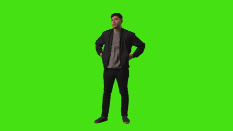full length shot of smiling casually dressed young man standing against green screen folding arms and putting hands in pockets