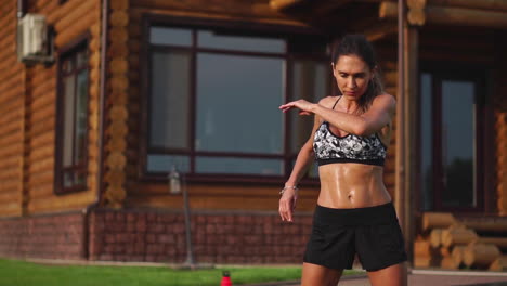 una mujer delgada y hermosa con ropa deportiva con una prensa abierta se está preparando para comenzar a entrenar en el césped cerca de su casa estirando sus brazos y hombros con ejercicios de calentamiento