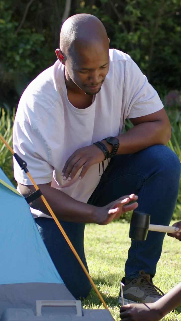 Vertical video: Guiding man in tee yard holding peg while child grabbing mallet hitting to fix tent