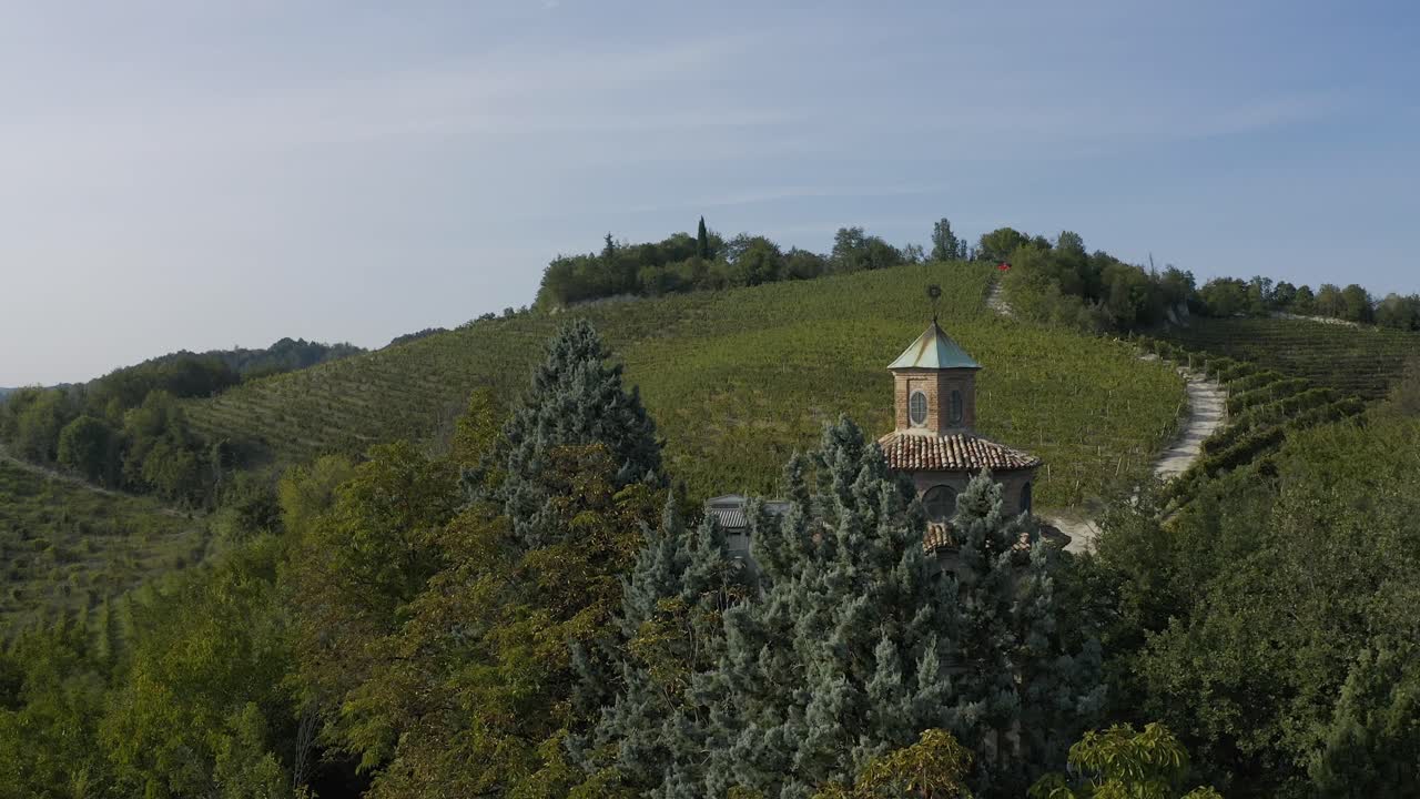 Vista aerea con drone della Cappella di San Giorgio su un fianco di collina, circondata da campi verdi e alberi, con un sentiero tortuoso che attraversa il paesaggio a Portofino sotto un cielo sereno.