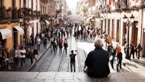 a lively urban environment filled with crowds walking along a bustling street. an older man sits peacefully, observing the energy of the busy city life around him.