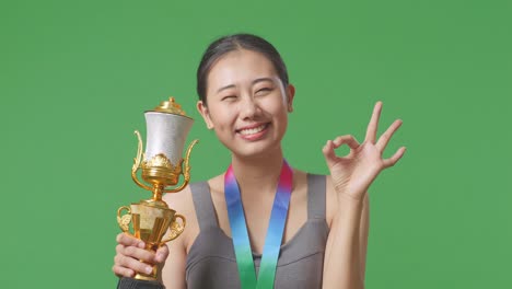 close up of asian woman with a gold medal and trophy showing okay gesture and smiling to camera as the first winner on green screen background in the studio