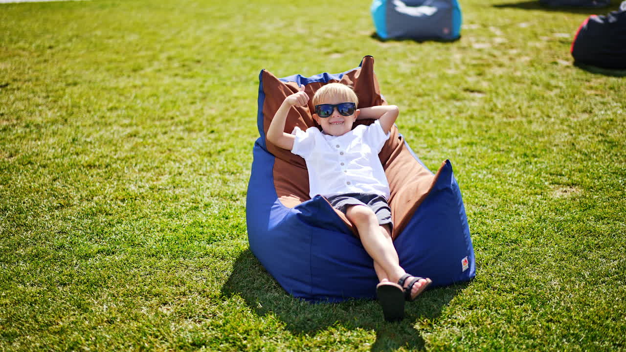 Smiling blond little boy enjoying his time in bean bag chair outdoors. Child in summer clothes and sunglasses relaxing and sunbathing.