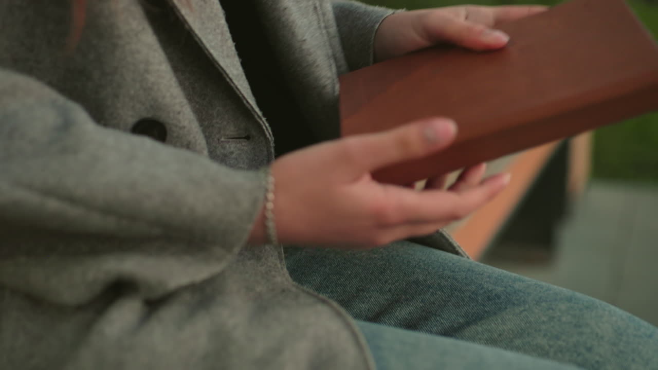 Primo piano di una donna seduta su una panchina di legno, con la mano appoggiata sul cappotto vicino al ginocchio, indossa un cappotto grigio e jeans di denim all'aperto, con particolare attenzione alla postura, alla texture e allo stile casual in un ambiente naturale
