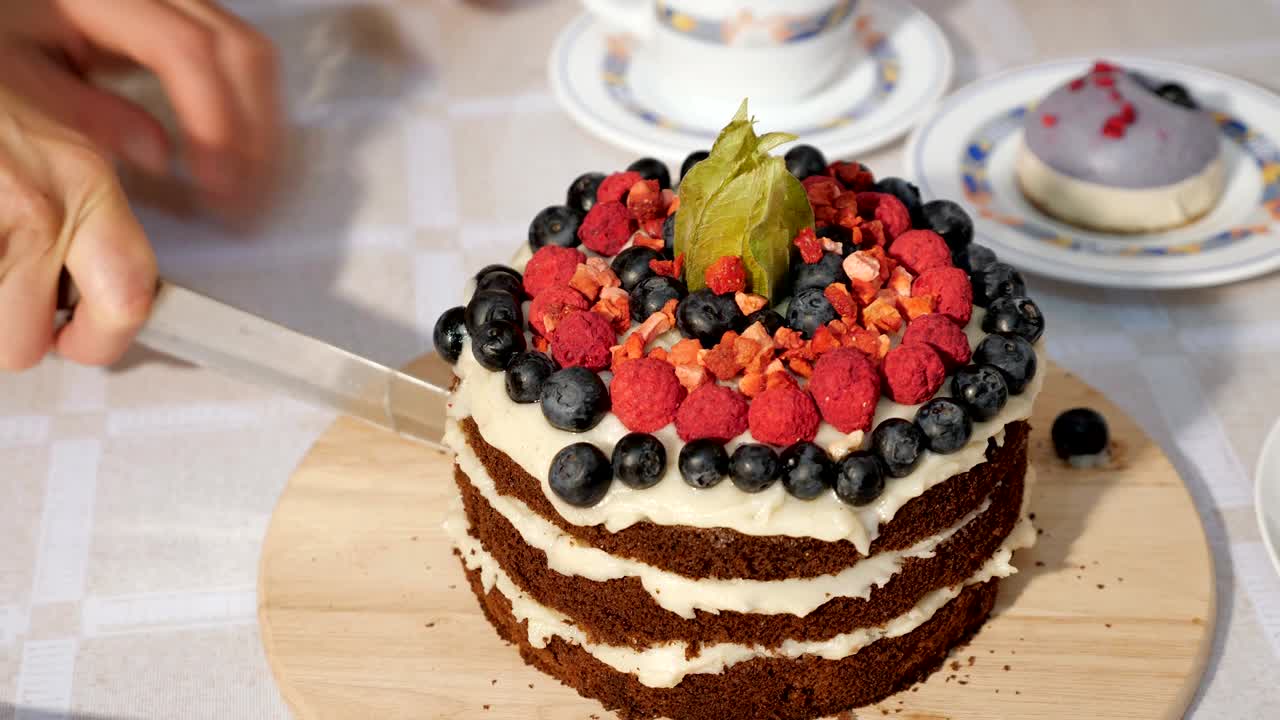 Donna taglia una fetta di torta di compleanno al cioccolato fatta in casa con strati di crema e pan di Spagna, decorata con frutti di bosco freschi