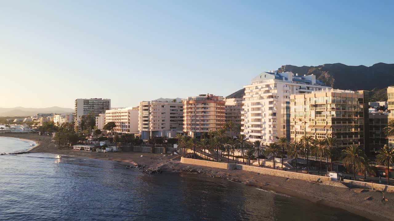 Splendida ripresa aerea che si allontana da un lussuoso edificio di Marbella di fronte alla spiaggia, con il riflesso del sole sul vetro che rivela la costa mediterranea fiancheggiata da palme durante l'ora d'oro.
