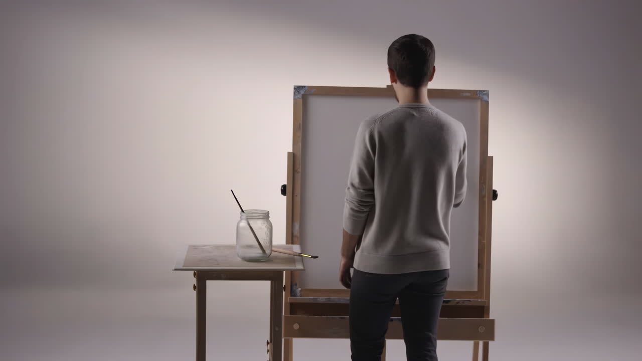 A male artist stands with his back to the viewer, facing a blank canvas on an easel in a studio