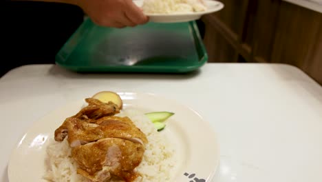 A person places plates of roasted and steamed Hainanese chicken rice on a counter under bright indoor lighting, with smooth camera movement and a clean dining environment