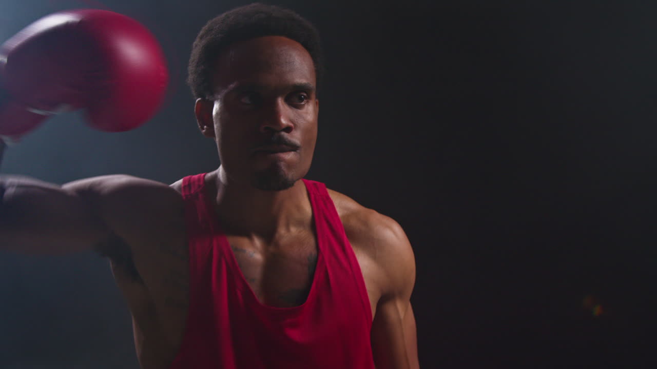 un atleta musculoso con una camiseta roja está de pie en un estudio oscuro, frente a la cámara con determinación y enfoque.