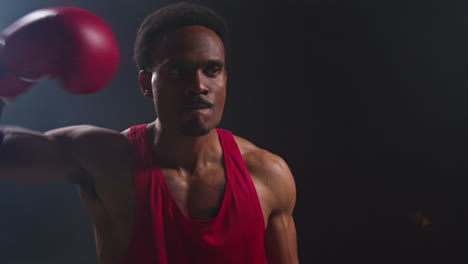 un atleta musculoso con una camiseta roja está de pie en un estudio oscuro, frente a la cámara con determinación y enfoque.