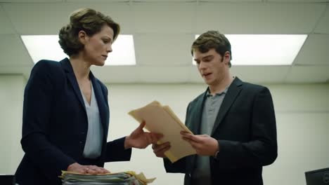 a heated discussion unfolds in a dimly lit office as a woman confronts a younger colleague about critical files. their expressions reflect stress and urgency while surrounded by paperwork.