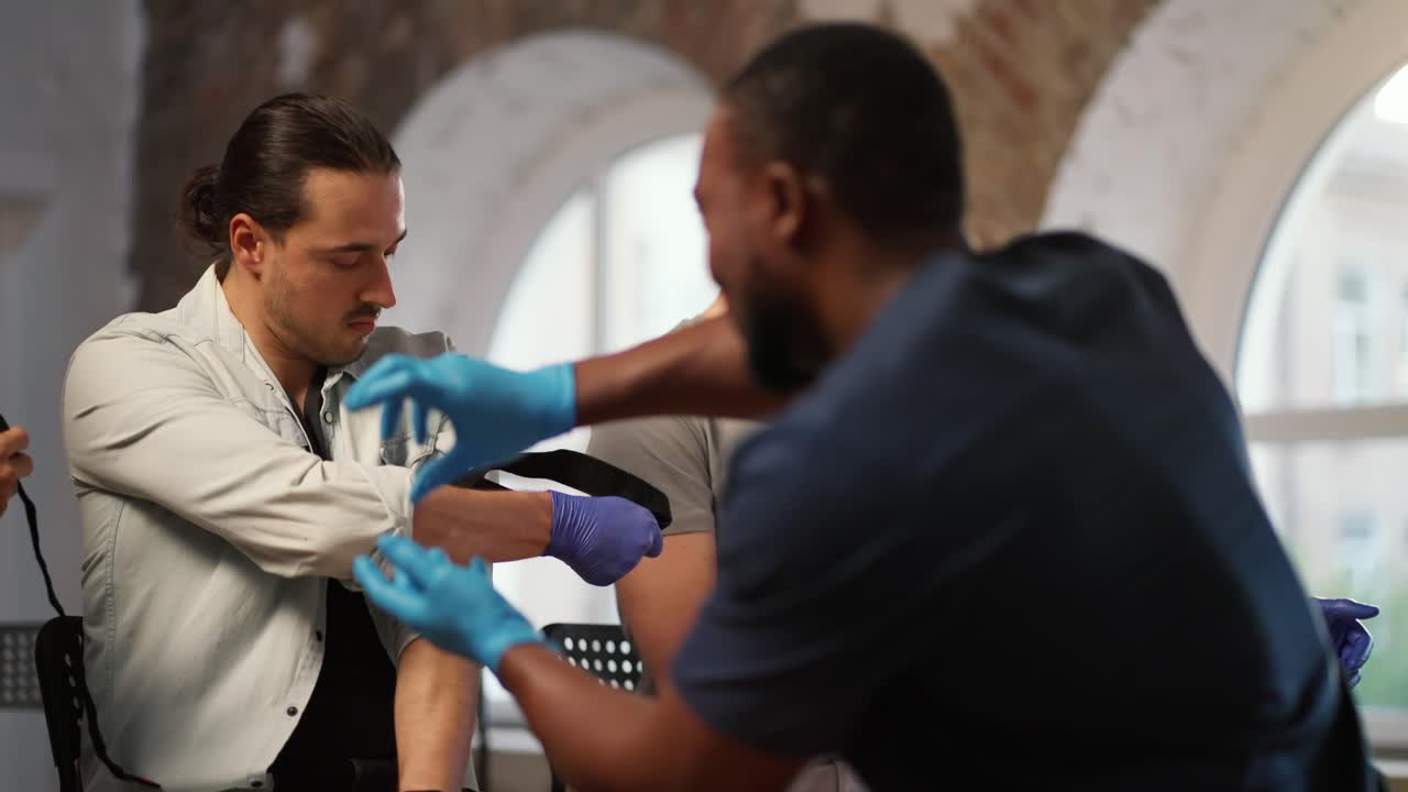A group of people perform practical exercises in medicine for first aid under the supervision of a professional black male doctor against the background of large windows. Course of a medic