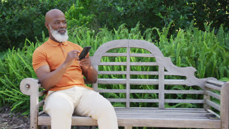hombre mayor afroamericano usando un teléfono inteligente mientras está sentado en un banco en el jardín