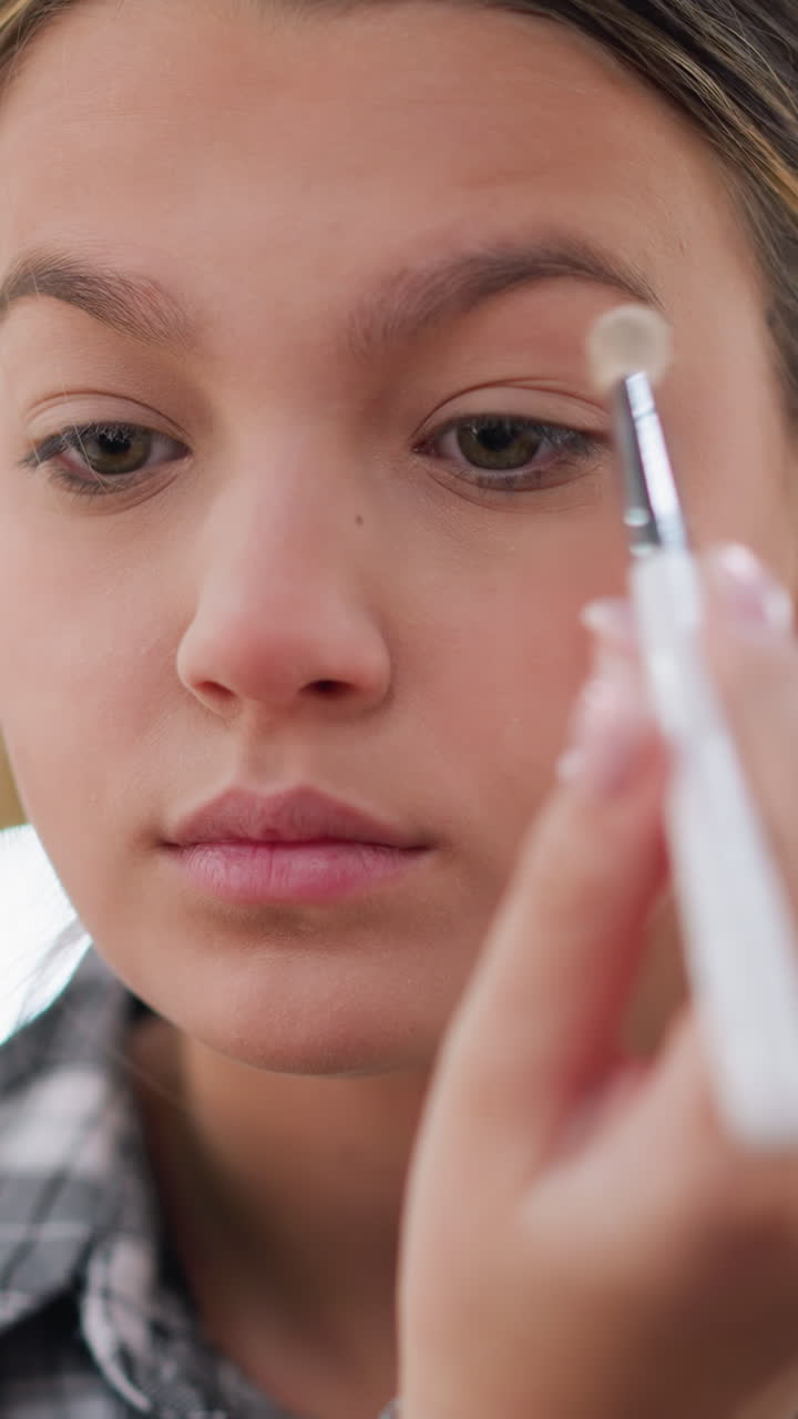 Adolescente maquillándose los ojos por la mañana con un pincel, primer plano suave de una chica joven creando un look natural para el día, tonos neutros suaves, expresión relajada, camisa de cuadros informal en casa
