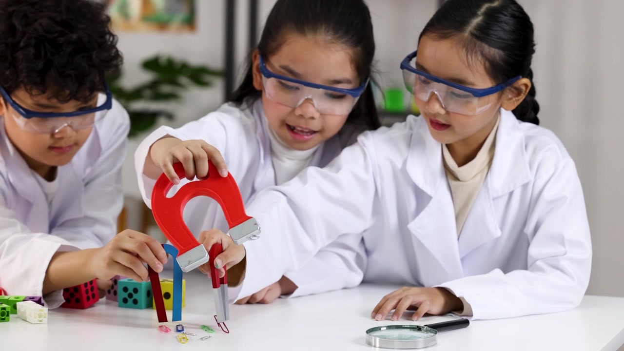 Asian children wear safety goggles and lab coats while testing magnets in a classroom.