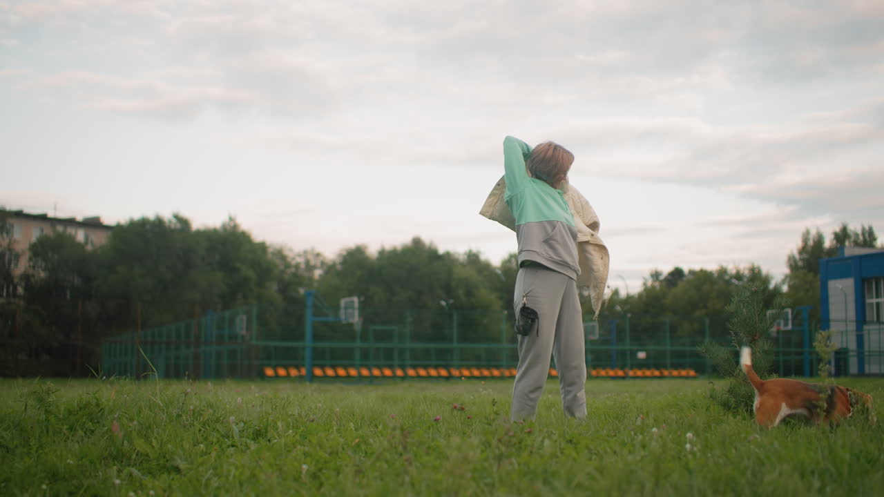 Elegant lady in hoodie after workout exercise, putting on hoodie as her playful puppy runs around in grassy outdoor park, enjoying fresh air after exercise session, bonding with her