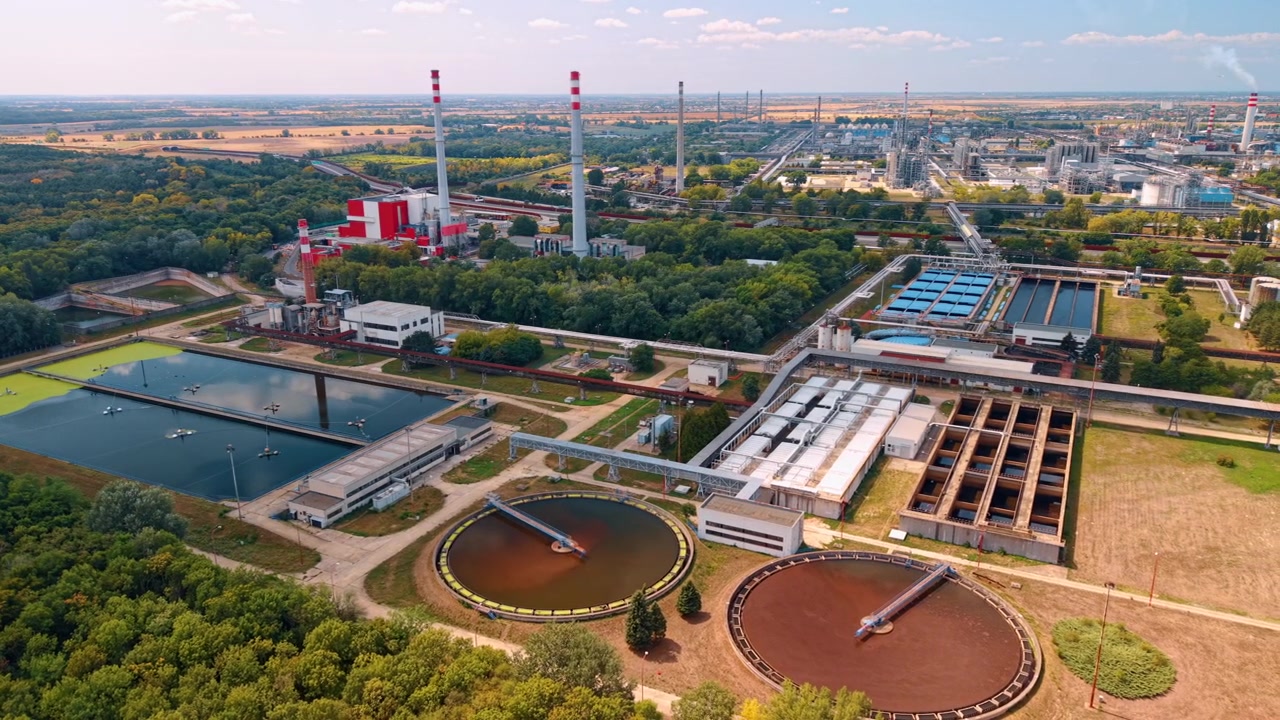 Wastewater clarifiers and treatment basins beside industrial stacks. Circular settling pools and rectangular channels fill the foreground below pipes, trees and chimneys.