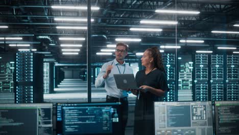 centro de datos mujer negra empresaria de negocios electrónicos y hombre blanco especialista en ti hablan, usan portátiles. ingeniero de tecnología de la información y administrador de sistemas trabajan en una gran granja de servidores de computación en nube.