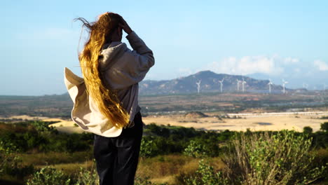 Woman-with-long-red-hairs-on-hilltop-watching-eco-friendly-wind-turbine-farm-in-background---Windy-day-in-Vietnam,Asia