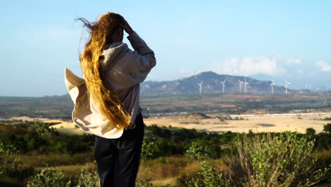 mujer con largos pelos rojos en la cima de una colina viendo una granja de turbinas eólicas ecológica en el fondo - día ventoso en vietnam, asia