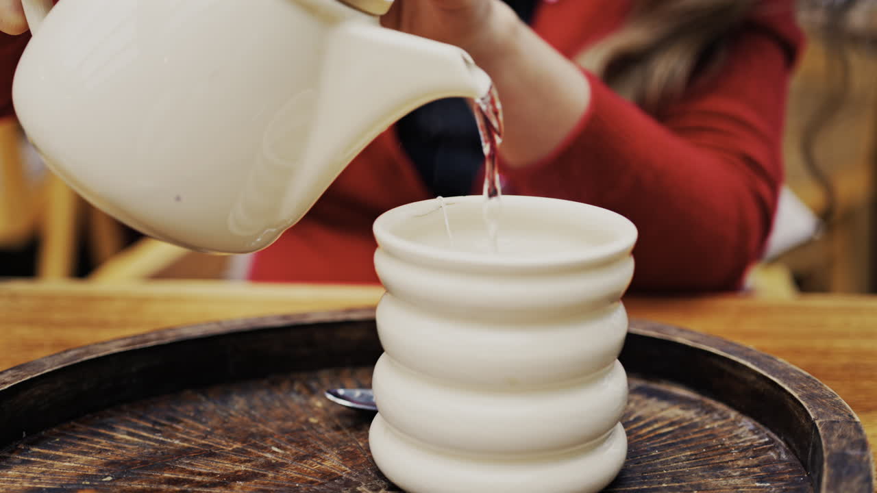 Close up of a woman wearing a red suit pouring hot water into a white cup with a tea bag in it on a wooden tray