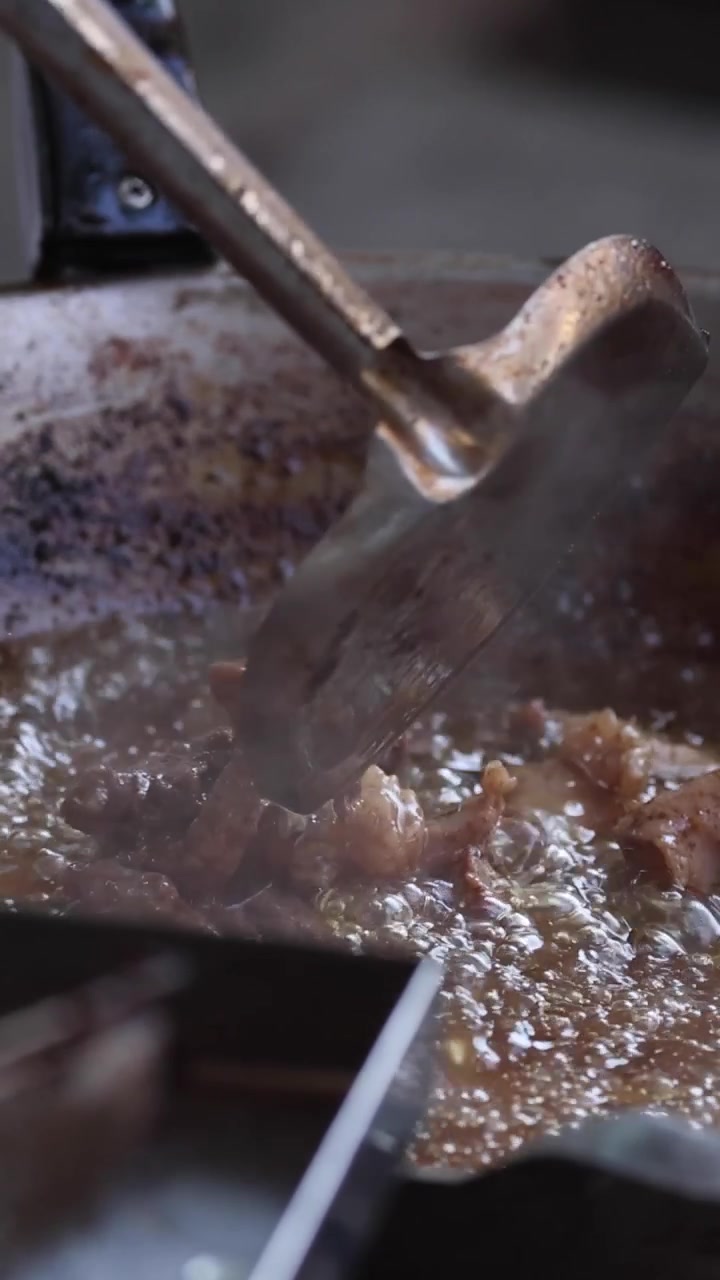 A metal spatula stirs pieces of meat in a bubbling, hot pan with rising steam.