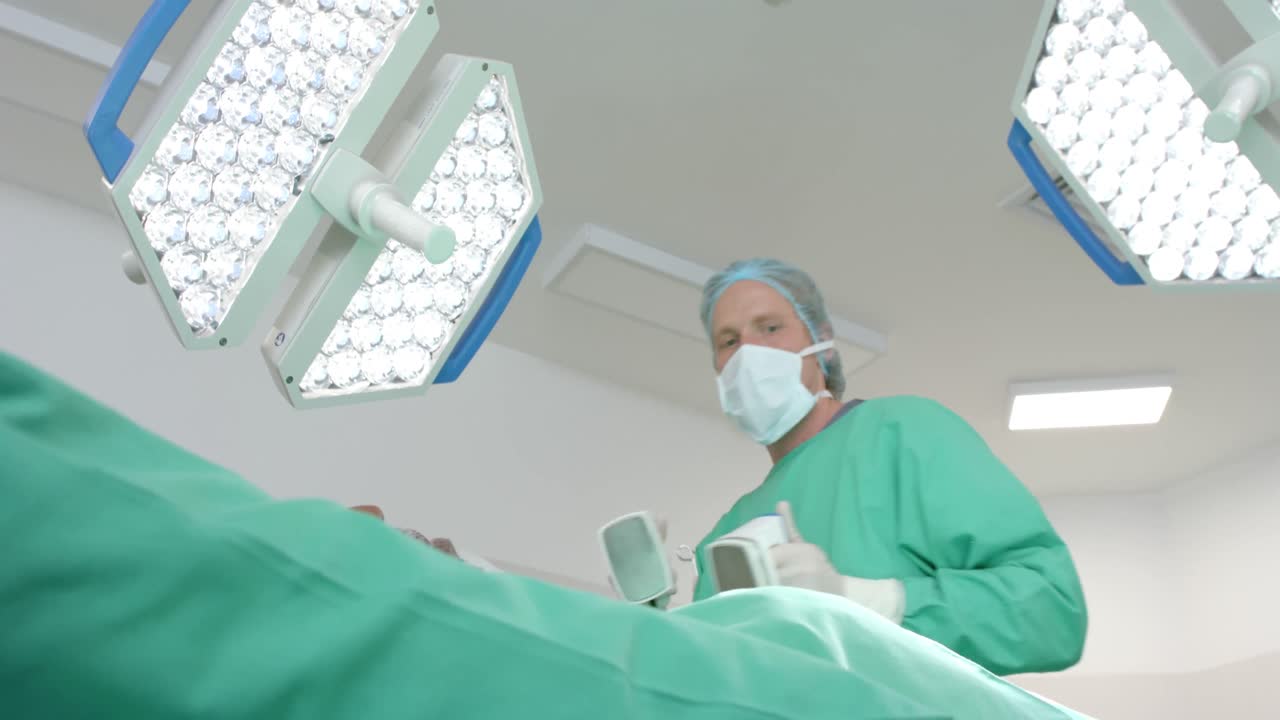 cirujano caucásico con bata quirúrgica, usando un desfibrilador en el paciente, cámara lenta