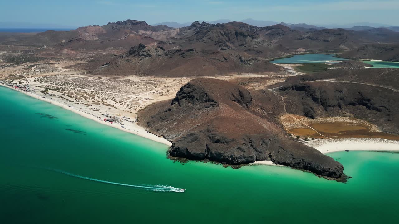 Paesaggio costiero con acque cristalline, una barca che sfreccia lungo la spiaggia e montagne in lontananza