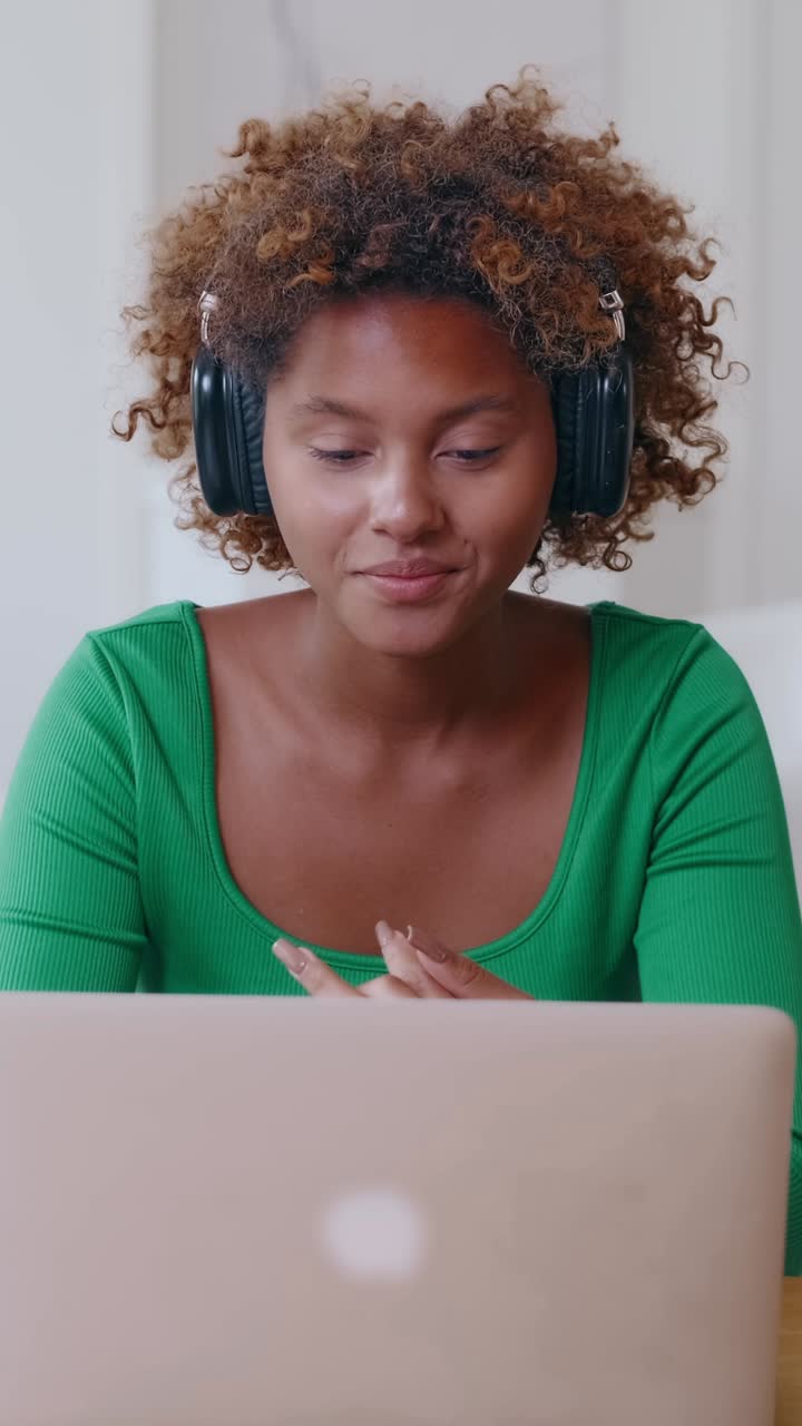 Young multicultural woman wearing headphones works on laptop in home office