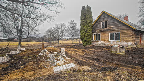 View-of-an-excavator-and-a-tractor-working-in-removing-all-the-rocks-and-levelling-the-land-for-gardening-besides-a-beautiful-wooden-cottage-on-a-sunny-day,-4K-in-timelapse