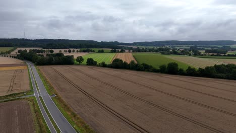 grey clouds at sky over agricultural farm fields in america. rural street between plantation and cultivated fields in summer. aerial wide shot. green hills in distance