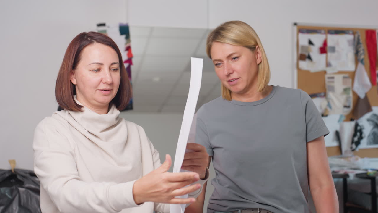 Tutor cheerfully explains paper pattern to student who lifts it up with focus, both women enjoy interaction in bright workspace surrounded by sewing materials, design board, and creativity tools
