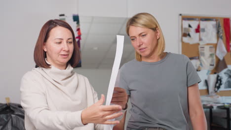 Tutor cheerfully explains paper pattern to student who lifts it up with focus, both women enjoy interaction in bright workspace surrounded by sewing materials, design board, and creativity tools