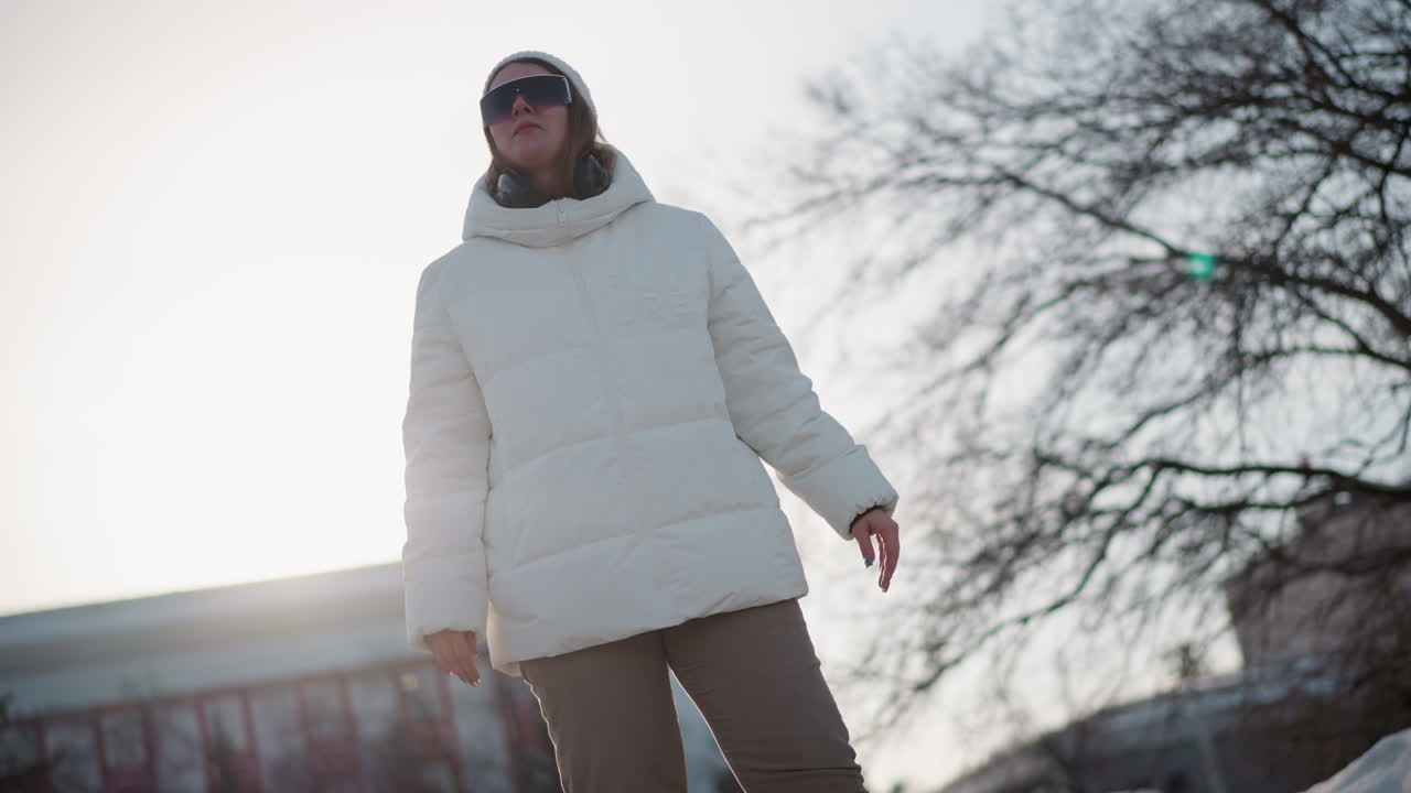 Elegant young woman standing under bright sun in snowy park wearing warm coat and beanie, adjusting tinted sunglasses while moving head to rhythmic beat, cheeks flushed by cold breeze