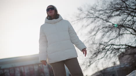 elegant young woman standing under bright sun in snowy park wearing warm coat and beanie, adjusting tinted sunglasses while moving head to rhythmic beat, cheeks flushed by cold breeze