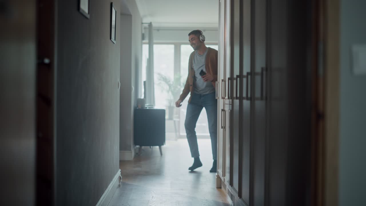 Portrait of a Happy Young Adult Man Listening to Music on Headphones Playing from His Smartphone at Home. Stylish Male in Casual Clothes is Carefree and Dancing in Corridor in His Apartment.