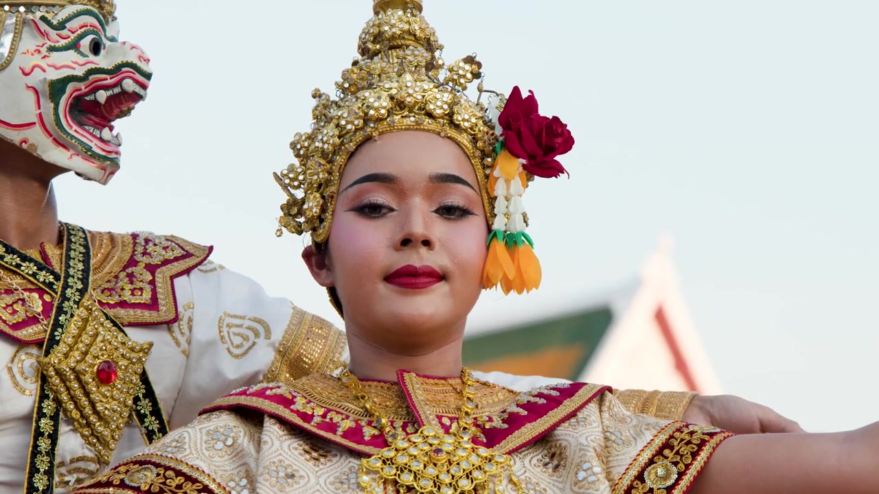 Performere i udsmykkede guldkostumer og masker danser udendørs ved et historisk thailandsk tempel.
