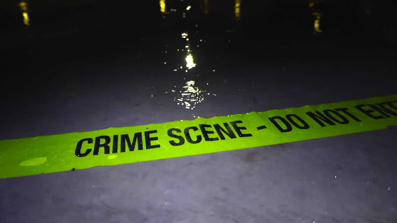 Yellow colorful and vibrant Crime Scene Do Not Enter tape thrown on a concrete slab where raindrops are falling in slow motion at night time. Well lit scene and light refelections on the water in back