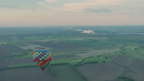 vibrant checkered hot air balloon drifting slowly over patchwork farmland, aerial perspective reveals patterned fields and distant horizon under soft pastel sky