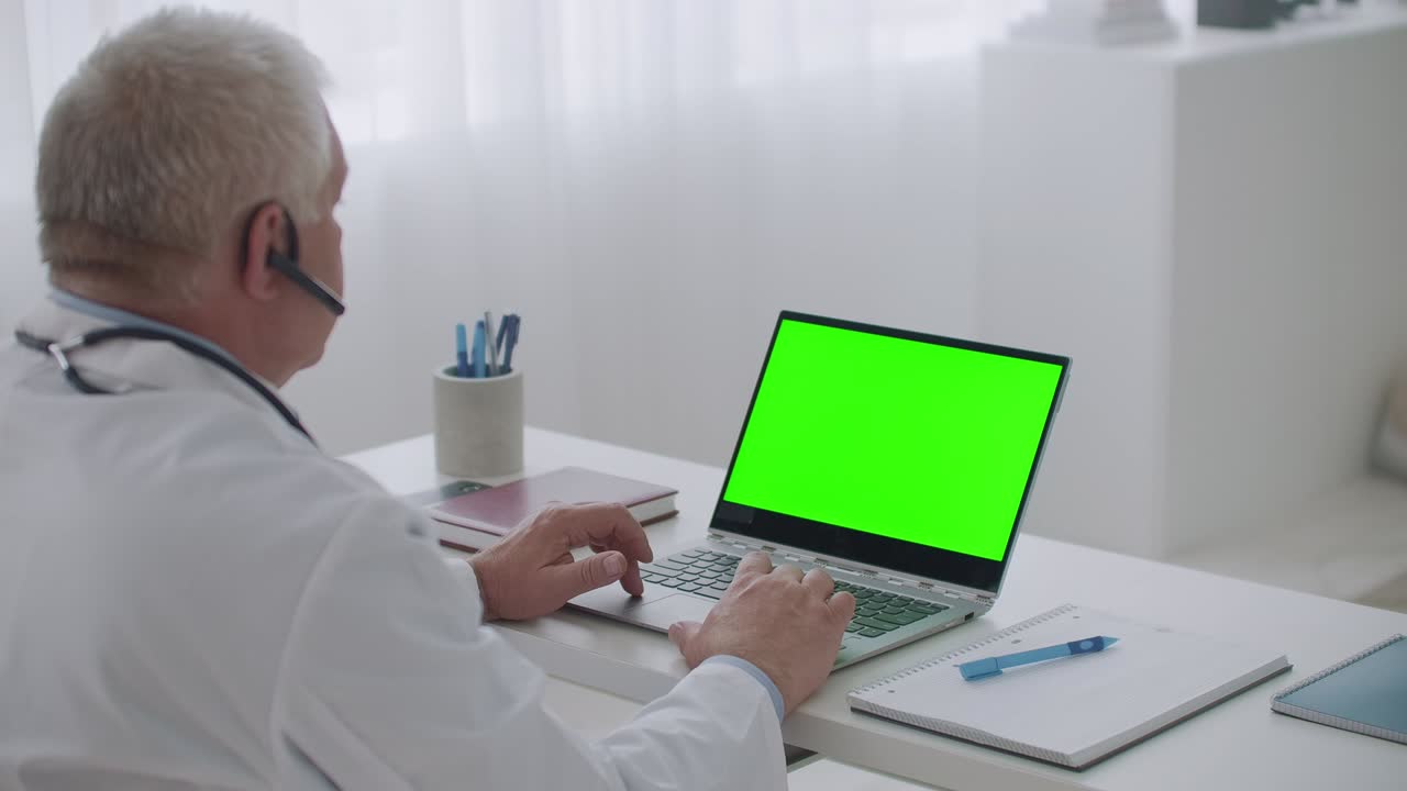 el médico experimentado está escuchando al paciente o colega por videollamada en una computadora portátil con pantalla verde para la tecnología de llave cromática