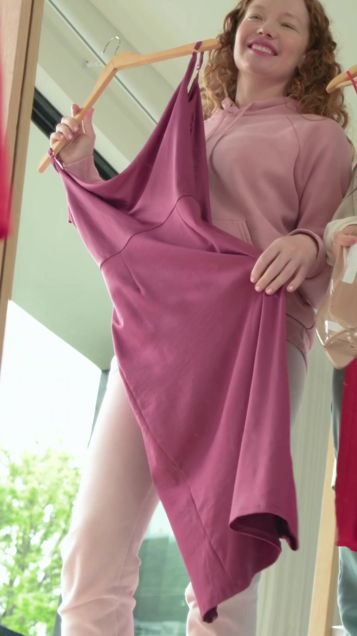 Video verticale: Donna con top rosa che raggiunge uno stendino, solleva un vestito color malva e ne controlla la caduta allo specchio.