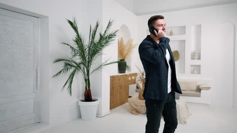 sportive caucasian man in jacket stands in the light modern room speaking on the phone. business conversation, talk with clients or partners.
